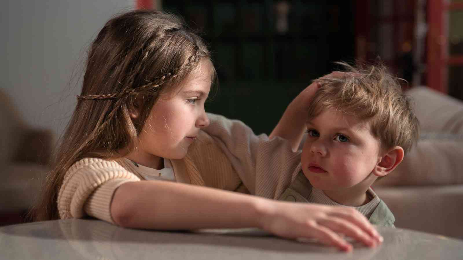 A young girl with braided hair gently places her hand on the head of a young boy. Both children are seated indoors, facing each other, with the boy looking at her intently.