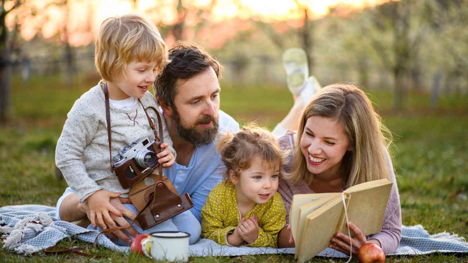 A family of four lies on a picnic blanket outdoors. The mother reads a book, the father observes, and one child holds a camera while the other looks on.