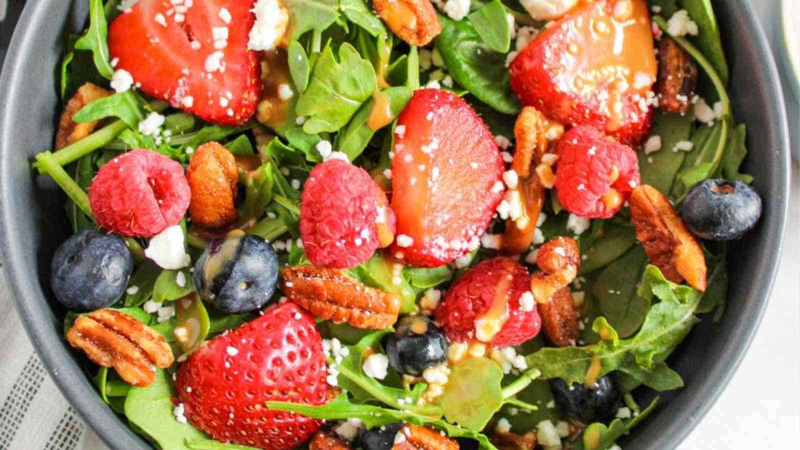 A salad in a black bowl containing leafy greens, strawberries, raspberries, blueberries, pecans, and crumbly cheese.