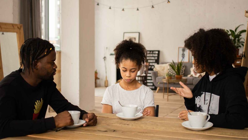 Three people at a table with white cups. A child sits between two adults, looking down. The adult on the right gestures with a hand. The setting is a well-lit room with a visible plant.