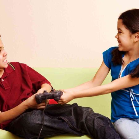 A boy and girl are playfully fighting over a game controller while sitting on a couch, both smiling.