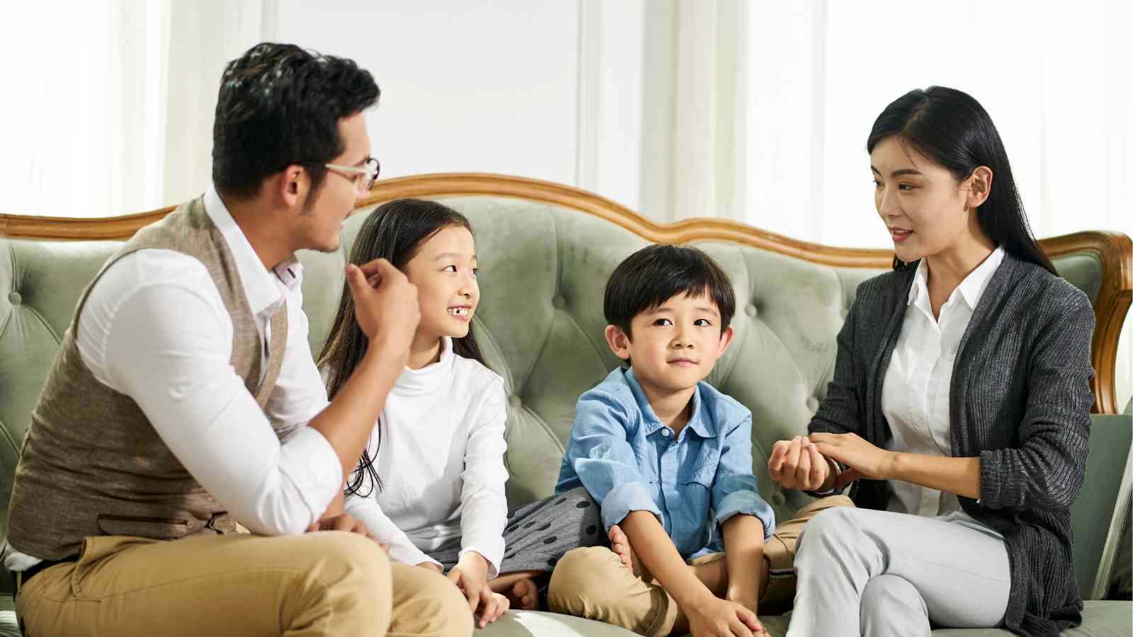 A family of four sits on a sofa, engaging in conversation. The father gestures as he speaks, while the mother and two children, a girl and a boy, listen attentively.