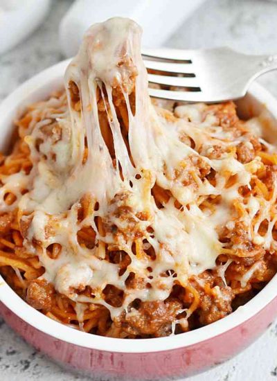 A bowl of baked spaghetti casserole topped with melted cheese, being lifted with a fork.