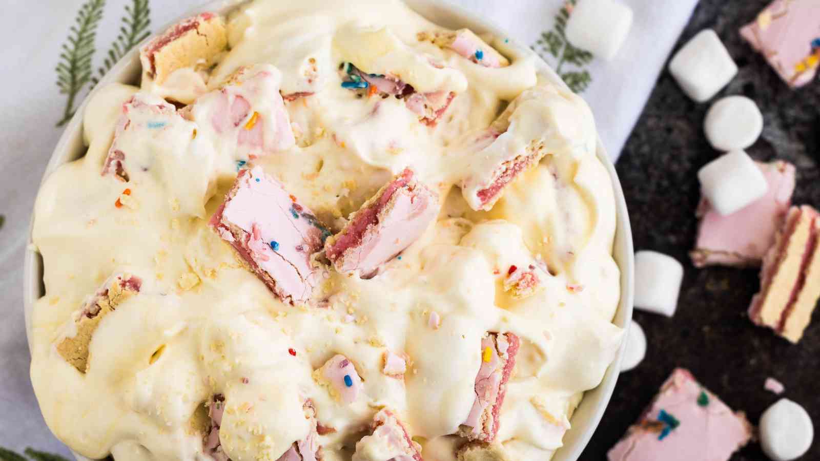 A bowl filled with creamy dessert topped with chunks of pink cookies, rainbow sprinkles, and small marshmallows on a white and black surface.