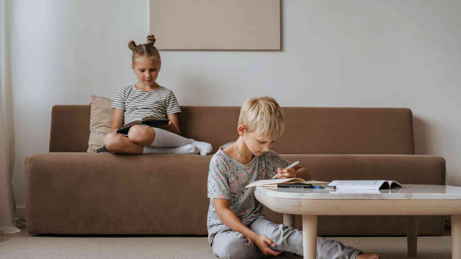 Two children in a living room: a boy sitting on the floor reading, and a girl on the couch using a tablet. A coffee table with books is in front of them.