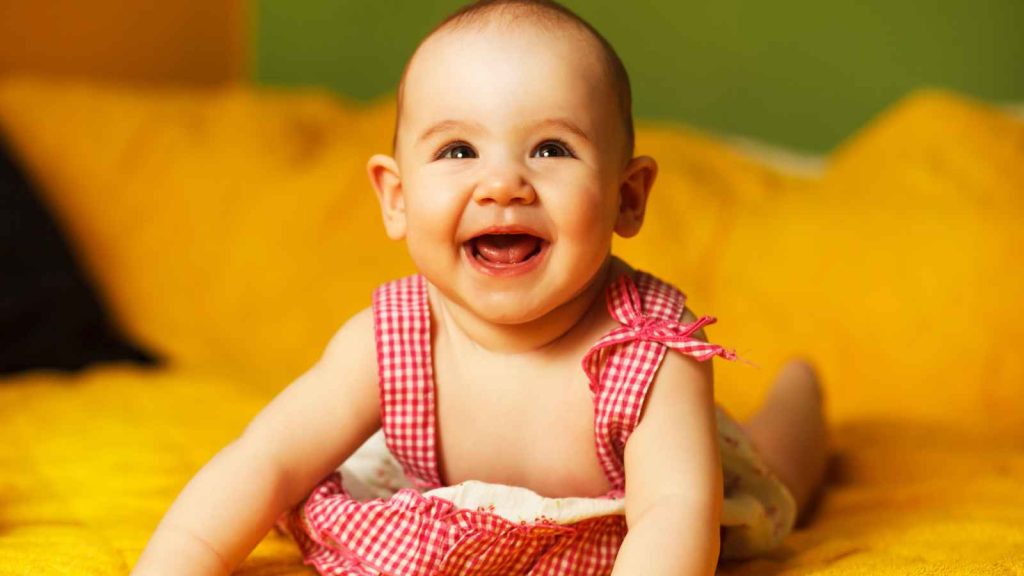 A smiling baby in a pink checkered outfit, lying on a yellow bedsheet.