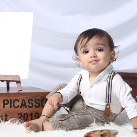A toddler in suspenders sits on fur beside a blank canvas on a mini easel labeled "Lil. Picasso Est. 2019," with a palette of paint in front.