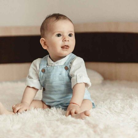 A baby wearing light blue overalls, sits on a fluffy white bed, gazing to the side with a curious expression.