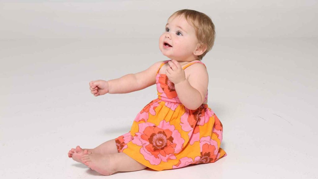 A baby wearing a bright floral dress sits on a white surface, looking upward with a curious expression.