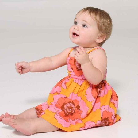A baby wearing a bright floral dress sits on a white surface, looking upward with a curious expression.