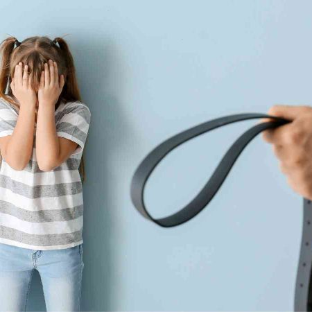A child covering her face stands against a wall, while an adult holds a belt in the foreground.