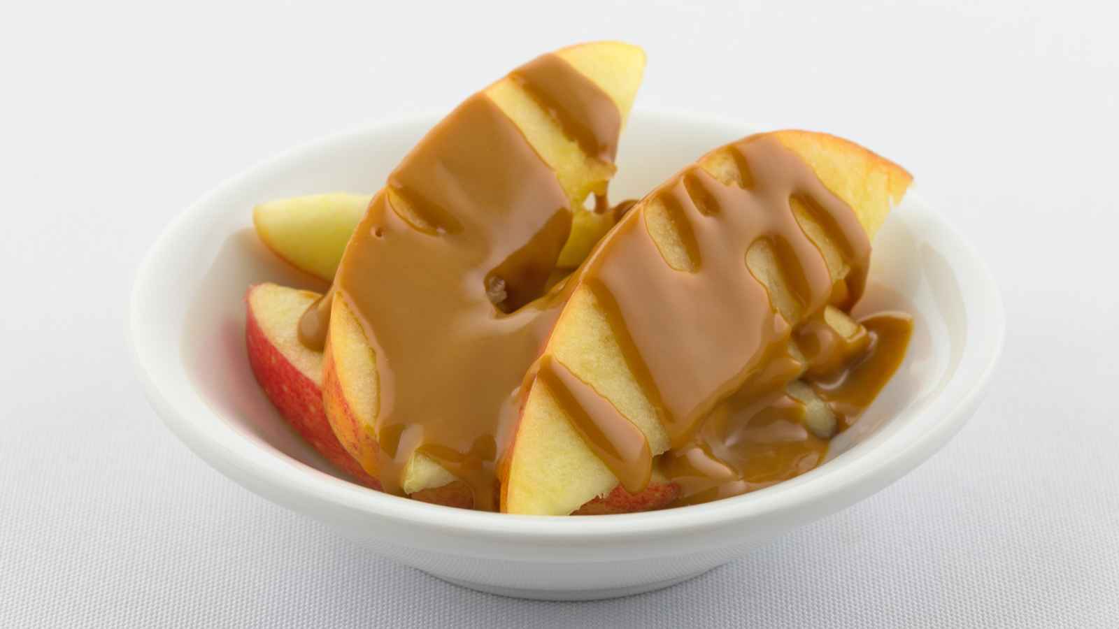 Apple slices drizzled with caramel sauce in a white bowl on a white surface.