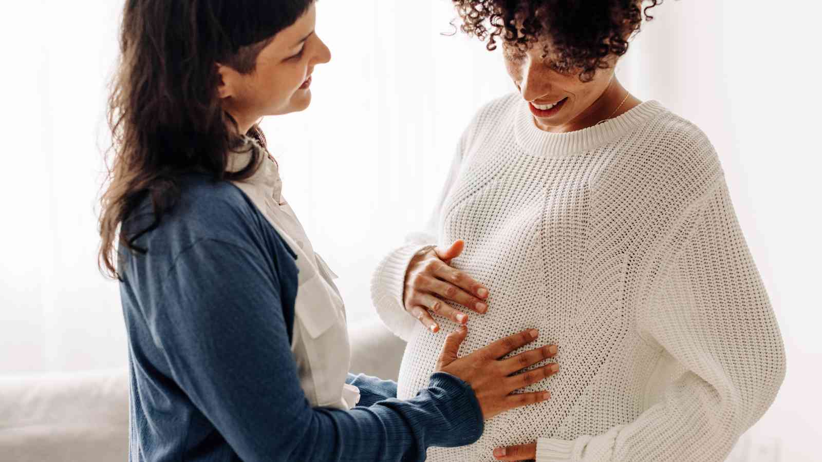 Two people smiling, with one person touching the other's pregnant belly. They are indoors, wearing casual outfits.