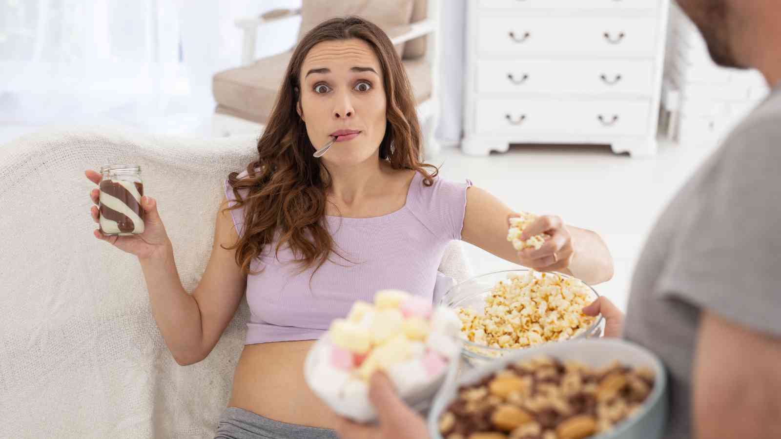 Pregnant woman on a couch eating popcorn and holding a jar with a spoon, while a person offers a bowl of snacks.