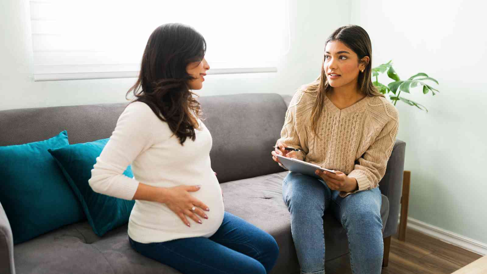 Pregnant woman in a white top sitting on a sofa, talking to another woman holding a clipboard. They are in a light-colored room with teal cushions and a plant in the background.