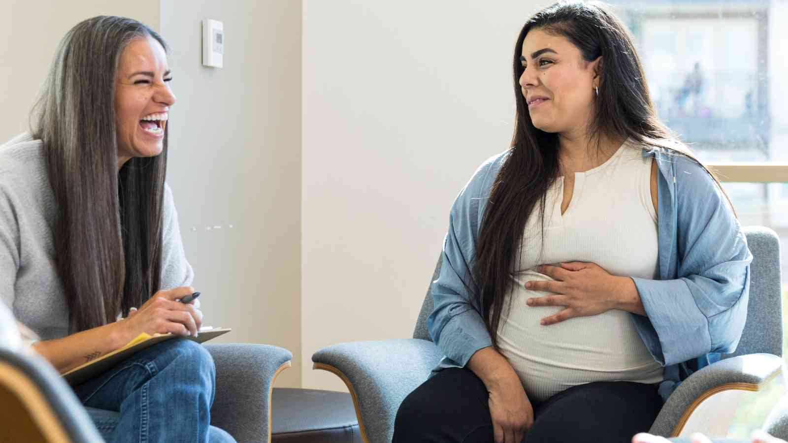 Two women sitting and talking in a bright room. One is holding a clipboard and laughing, while the other, visibly pregnant, touches her belly and smiles.