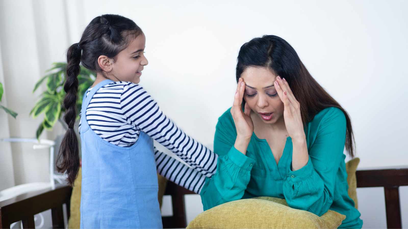 A woman sitting and holding her head in distress while a young girl stands beside her, smiling and placing a hand on her shoulder.