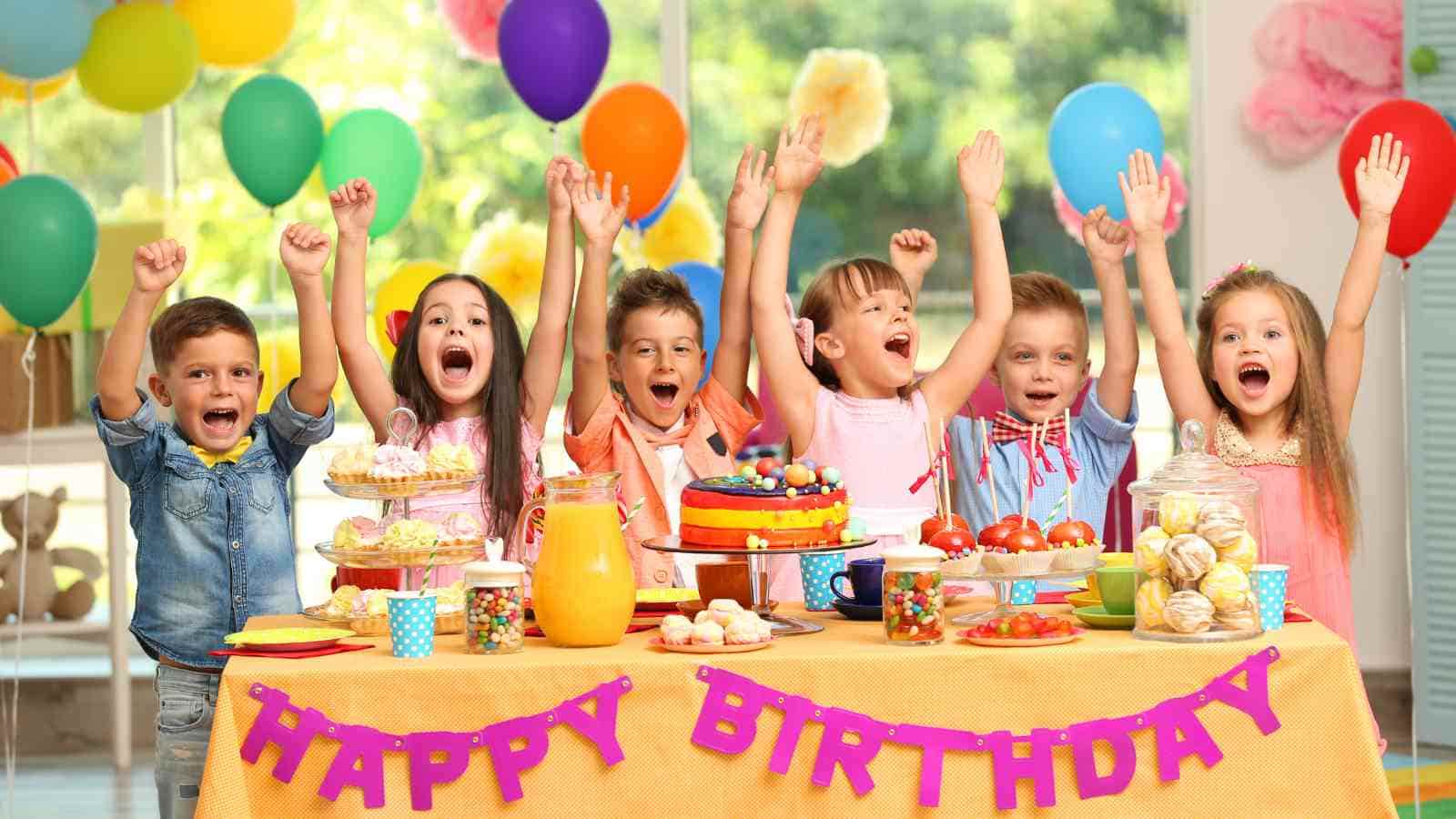 Seven children celebrating a birthday in front of a table with a cake, snacks, and drinks. Colorful balloons are in the background, and a "Happy Birthday" banner is visible.