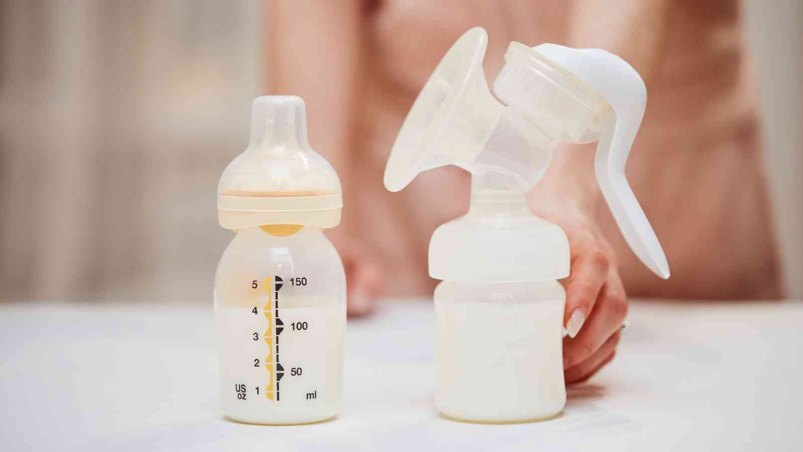 Breast pump and baby bottle with milk on a table, with a person in the background.