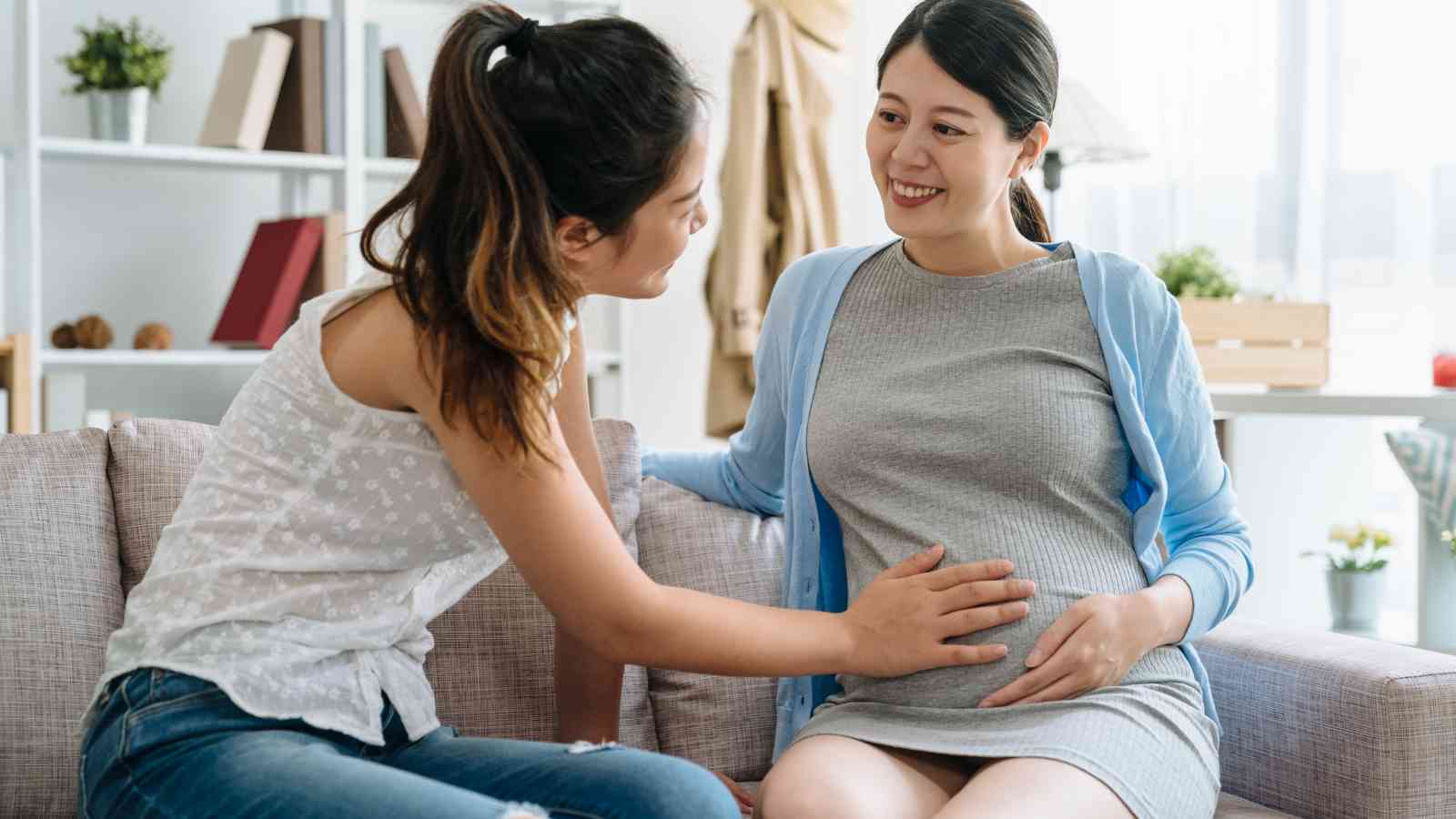A pregnant woman sits on a couch, smiling, while another woman touches her belly affectionately. Bookshelves are visible in the background.