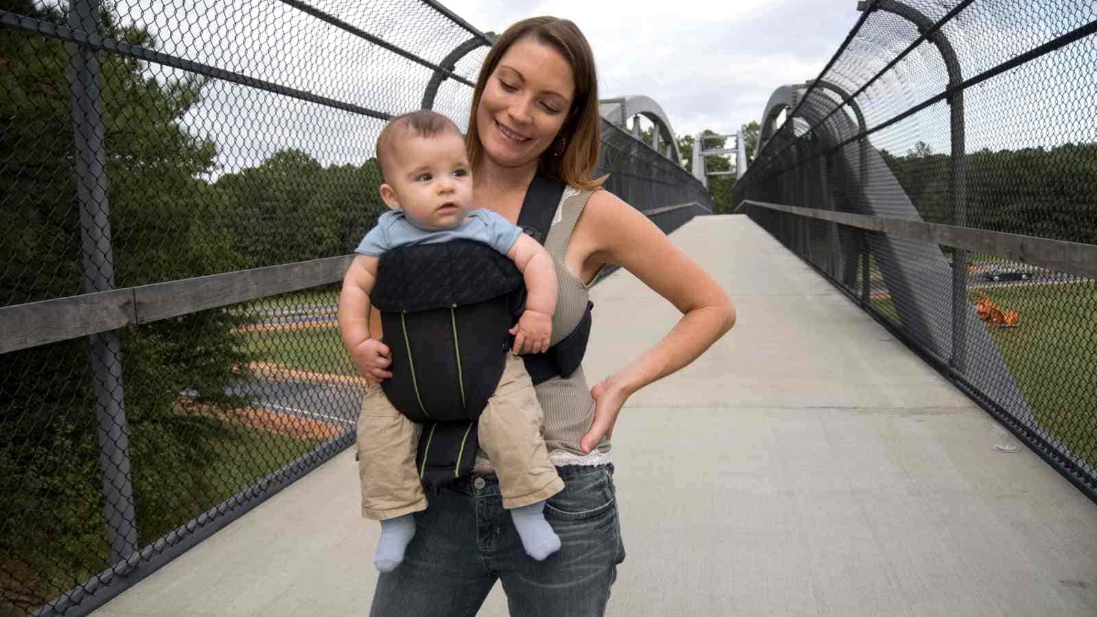 A woman stands on a bridge, smiling, while carrying a baby in a front-facing carrier.