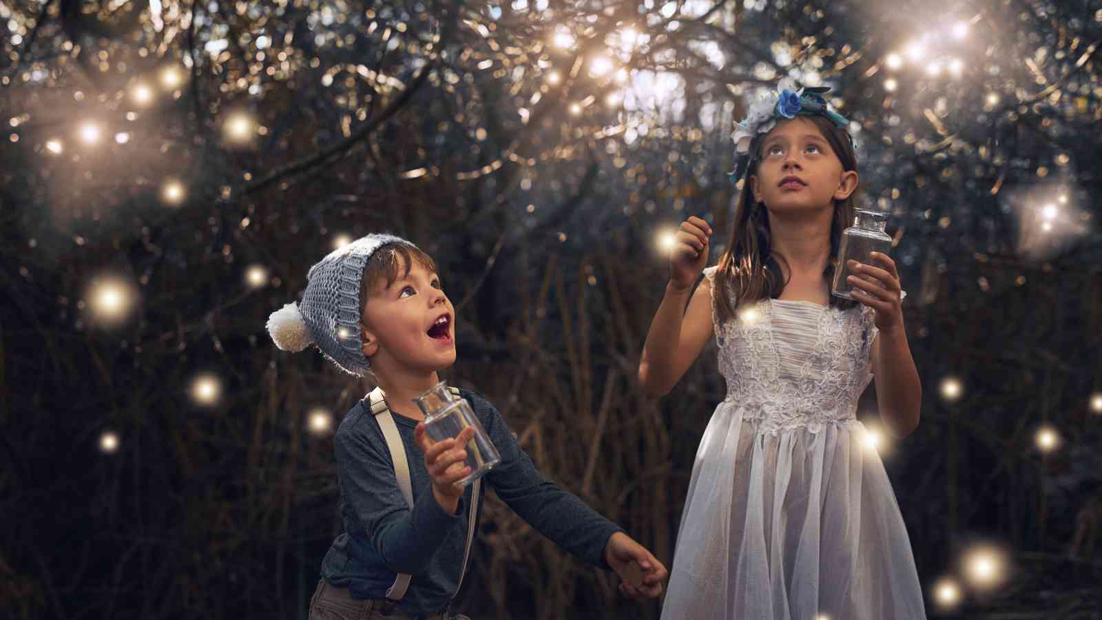 Two children stand in a forest, each holding a jar, surrounded by glowing lights. One child wears a knit hat, and the other a floral headband and white dress.