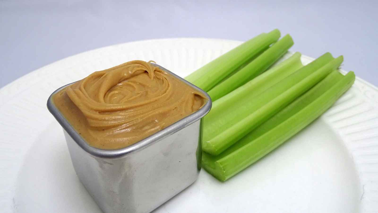 A small metal container of peanut butter next to several celery sticks on a white plate.