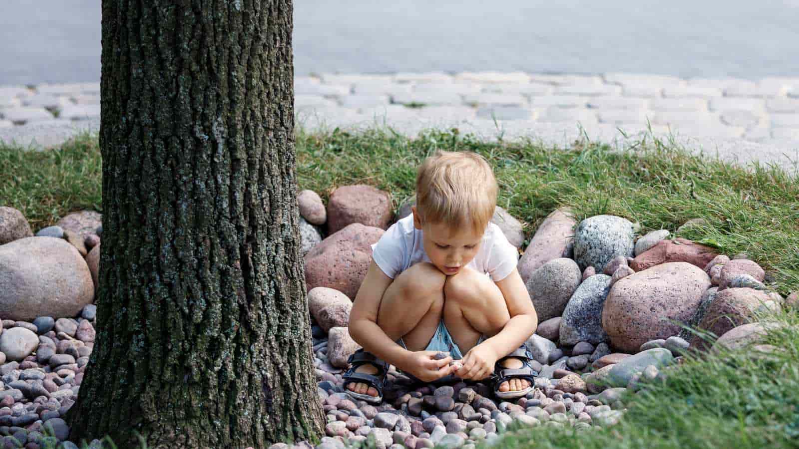 A child in a white shirt and sandals crouches on a bed of stones next to a tree trunk, intently looking at the ground.