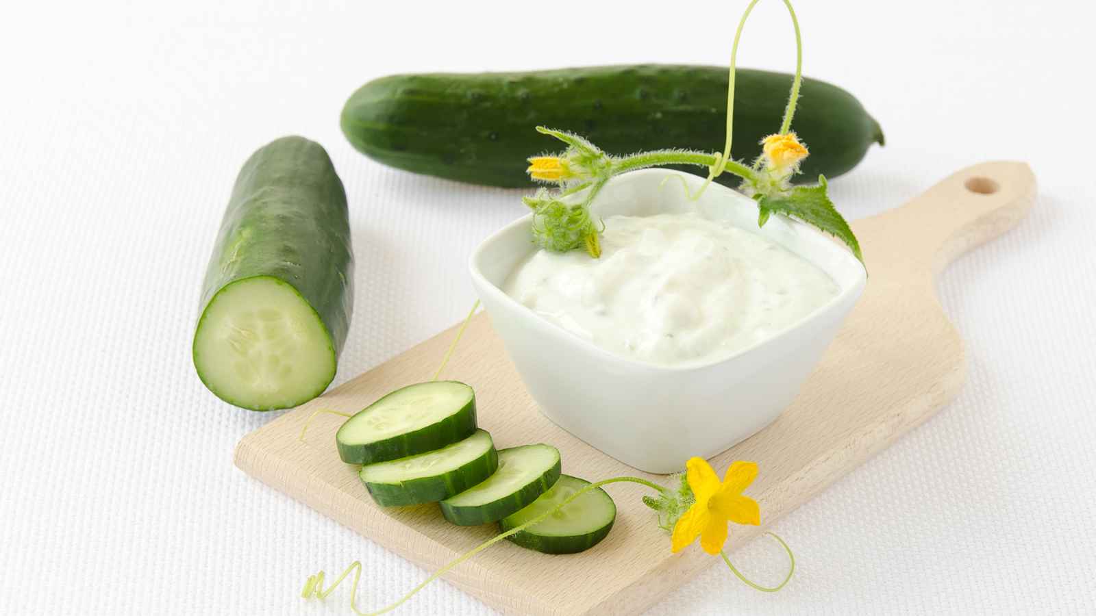 Cucumber slices and a whole cucumber beside a small bowl of cream, garnished with a yellow flower and tendrils, on a wooden board.