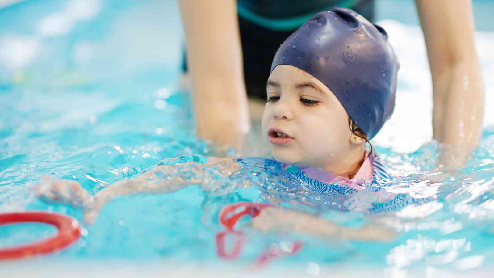 Child wearing a swim cap learning to swim in a pool, guided by an adult.