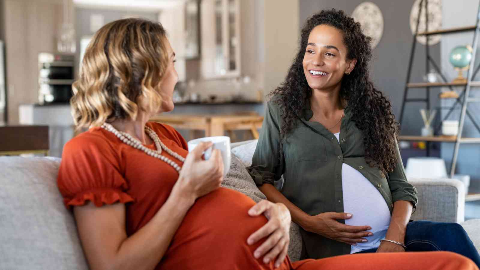 Two pregnant women sit on a sofa, smiling and talking. One holds a coffee cup, while the other rests her hand on her belly. The room is bright with modern decor.