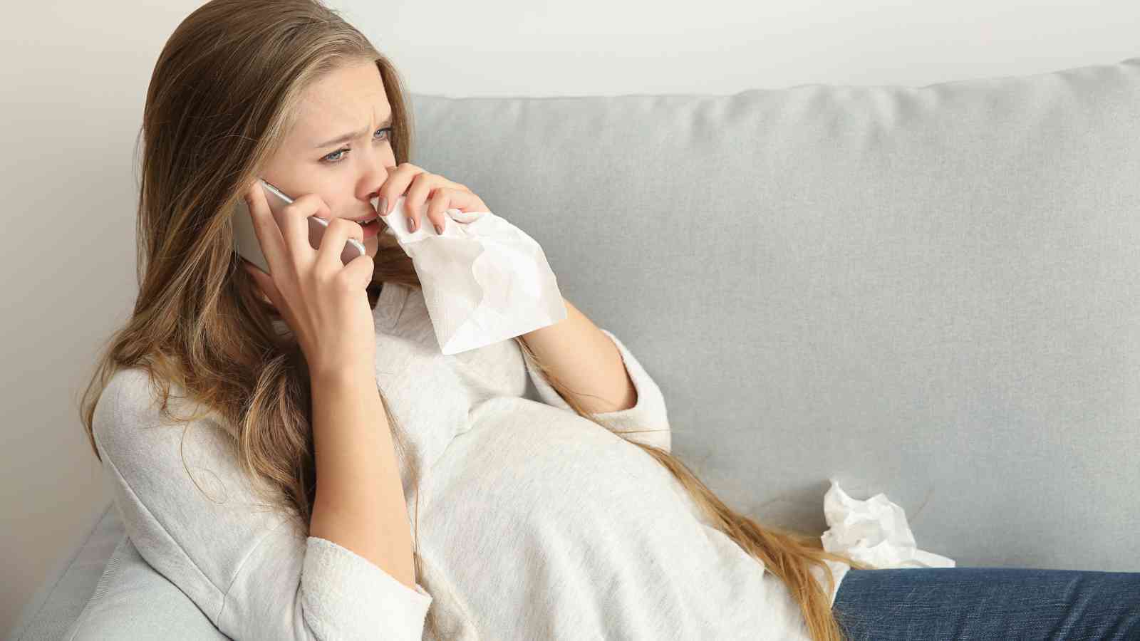 Pregnant woman sitting on a couch, holding a phone to her ear and a tissue to her face. She appears to be in distress or crying.