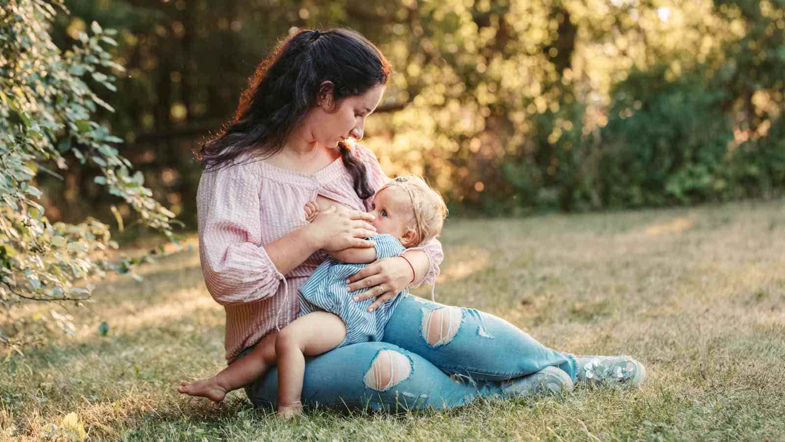 A woman sits on the grass breastfeeding a baby, surrounded by trees. She wears a light pink top and ripped jeans. The baby is in a blue outfit.