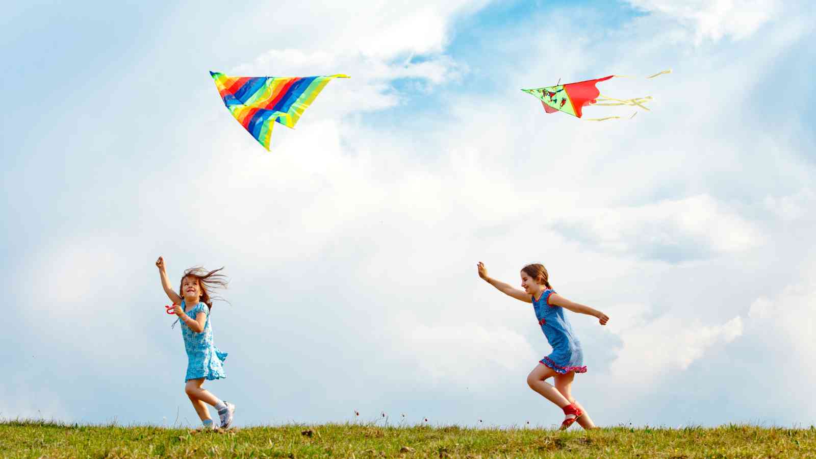 Two children running in a grassy field flying colorful kites against a cloudy sky.