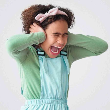 A child with curly hair covers their ears and has a distressed expression. They are wearing a green top and blue overalls, and a pink headband. The background is plain gray.