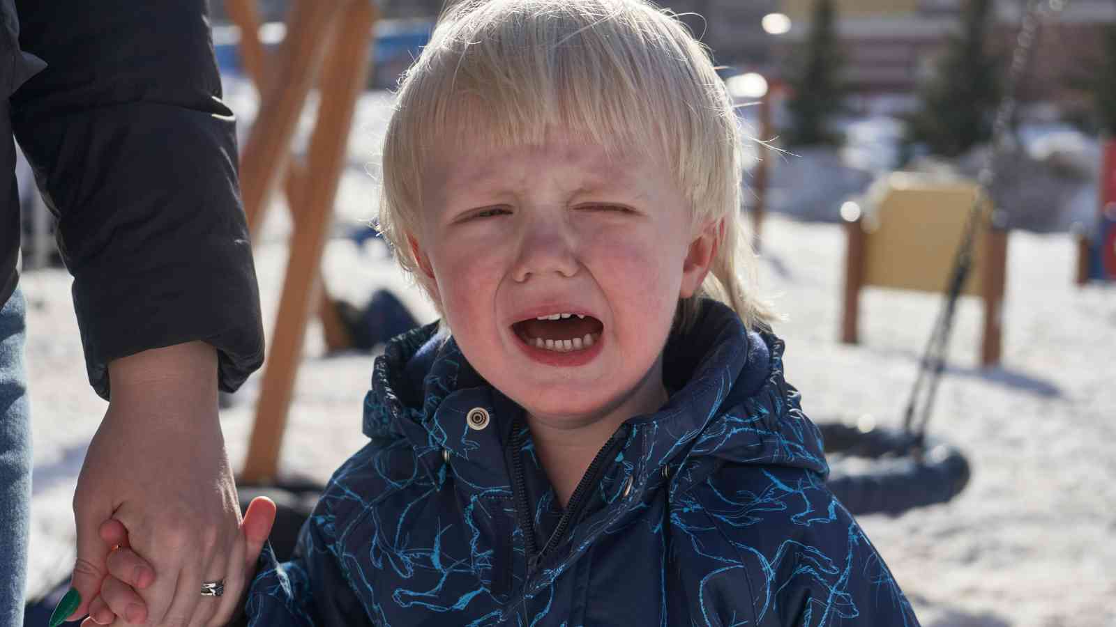 Child in a blue jacket crying outdoors, holding an adult's hand, with playground equipment and snowy background.