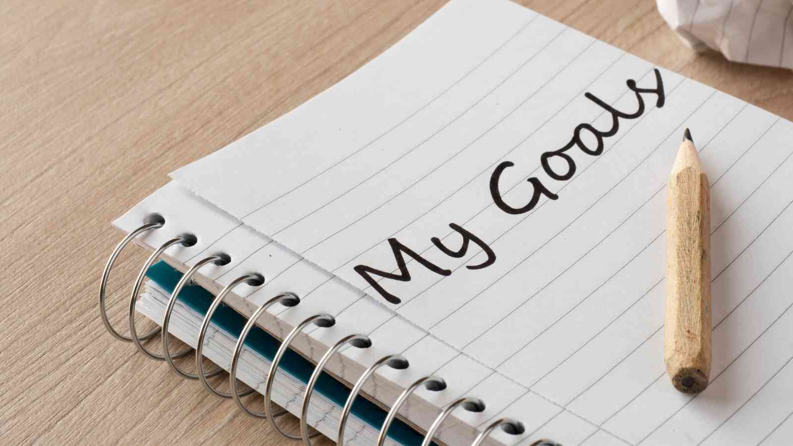 Open spiral notebook with "My Goals" written on a lined page. A sharpened wooden pencil rests on the right.