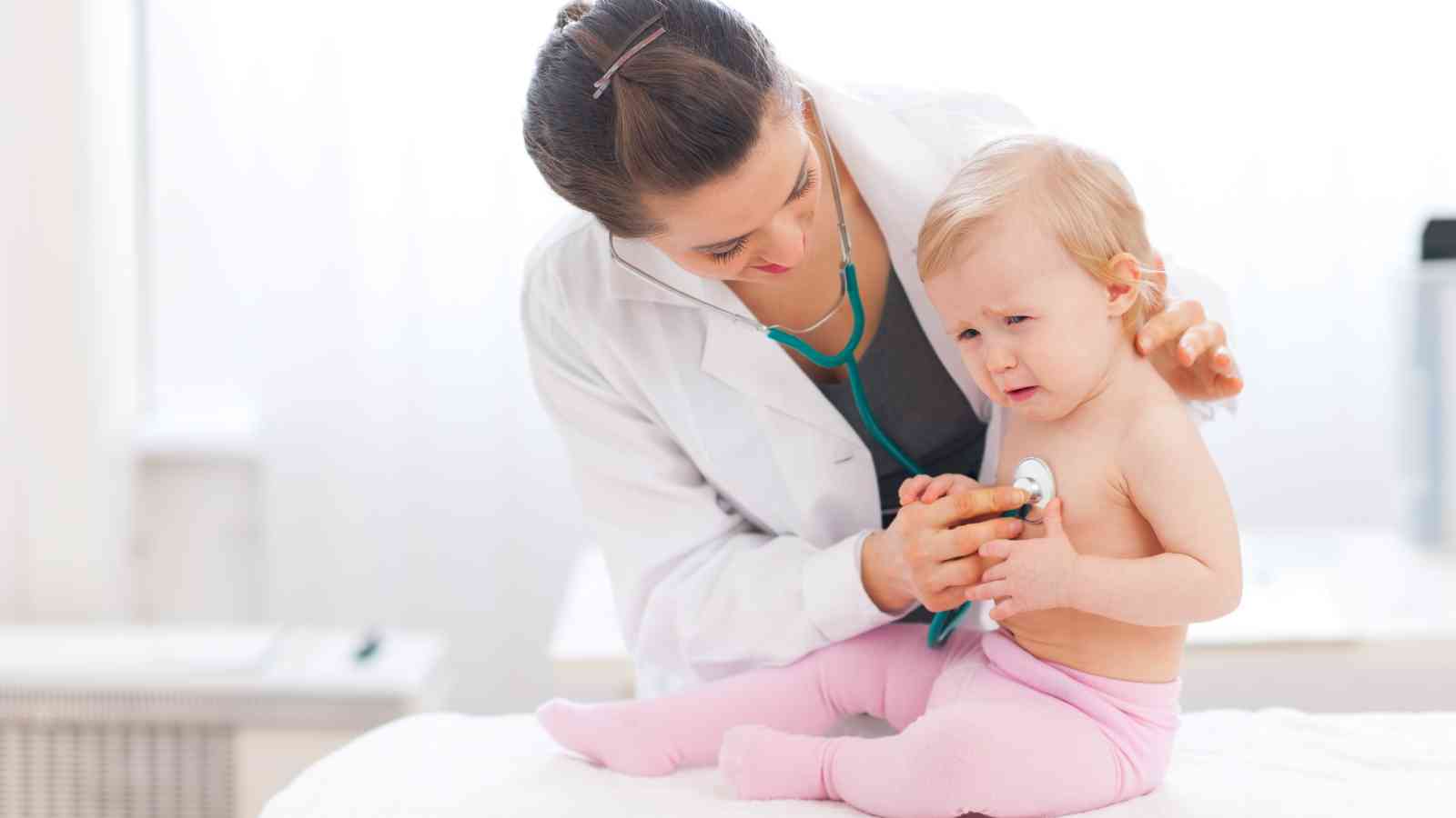 A pediatrician uses a stethoscope to examine a baby sitting on a table, who appears slightly upset.
