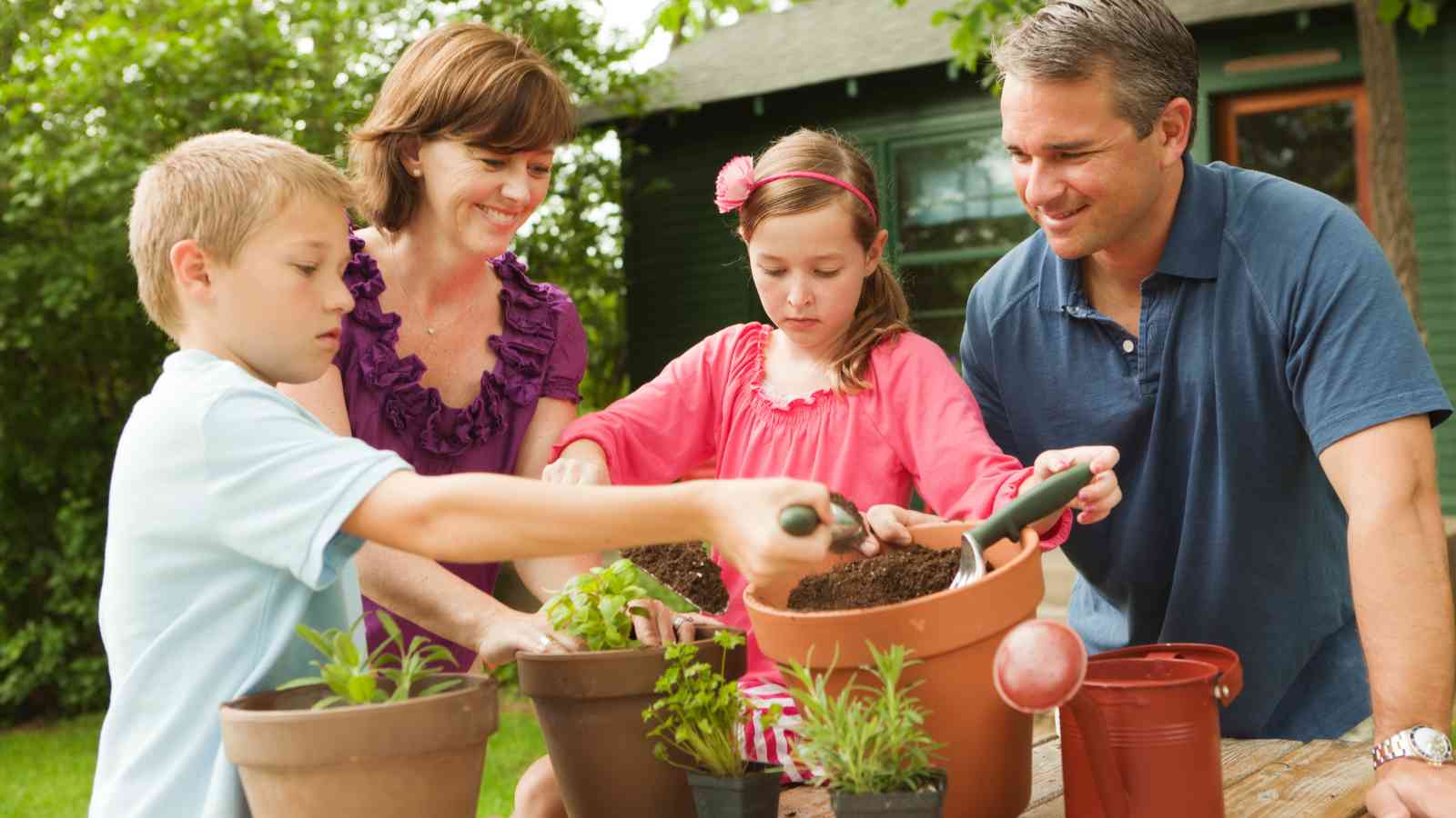 A family of four is gardening outdoors. The children are planting in pots, assisted by their parents. They're surrounded by greenery, with a house in the background.