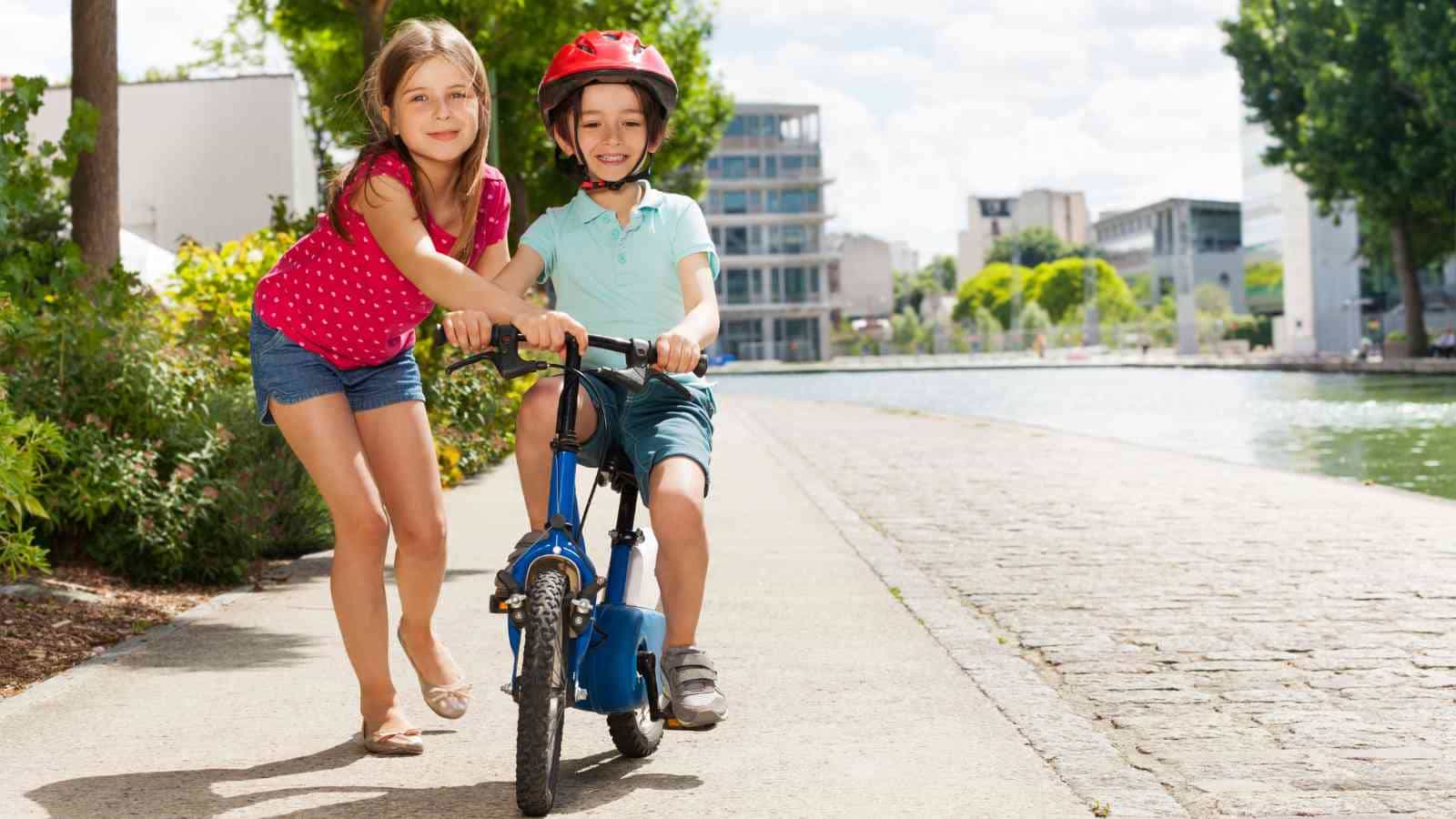 A girl helps a boy in a red helmet ride a blue bicycle on a sunny path by the water.