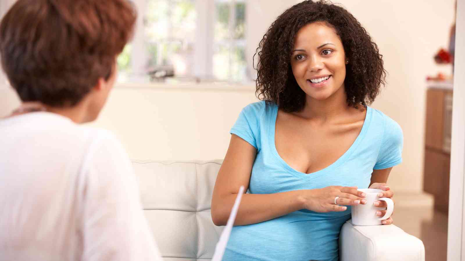 A woman with curly hair holds a mug and smiles while seated on a couch, conversing with another woman facing away.