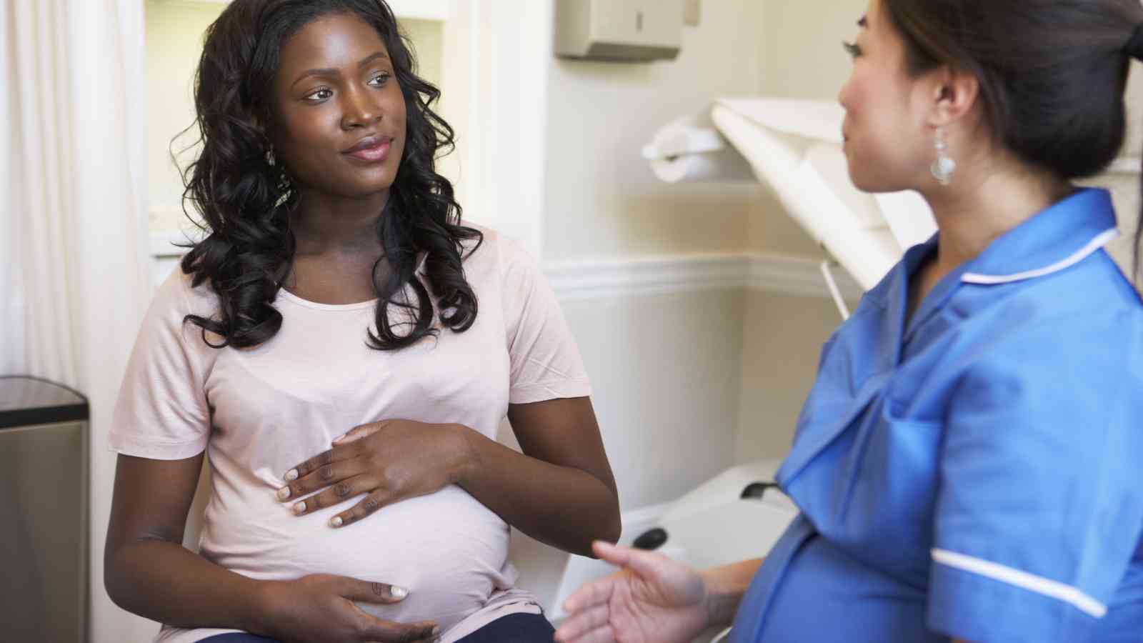A pregnant woman sits and talks with a healthcare professional in blue scrubs, in what appears to be a medical setting.