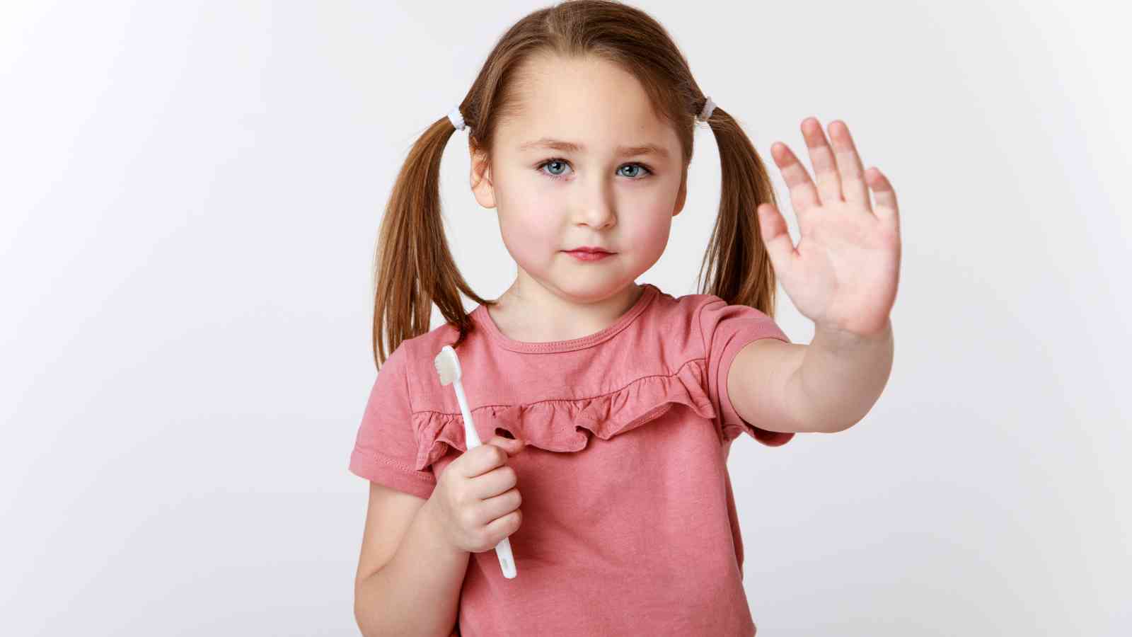 A young girl with pigtails holds a toothbrush in one hand and raises her other hand, palm open. She wears a pink shirt and stands against a plain white background.