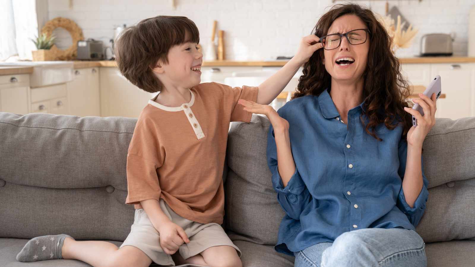 A boy playfully pulls a woman's hair as she winces while sitting on a gray couch in a kitchen setting.