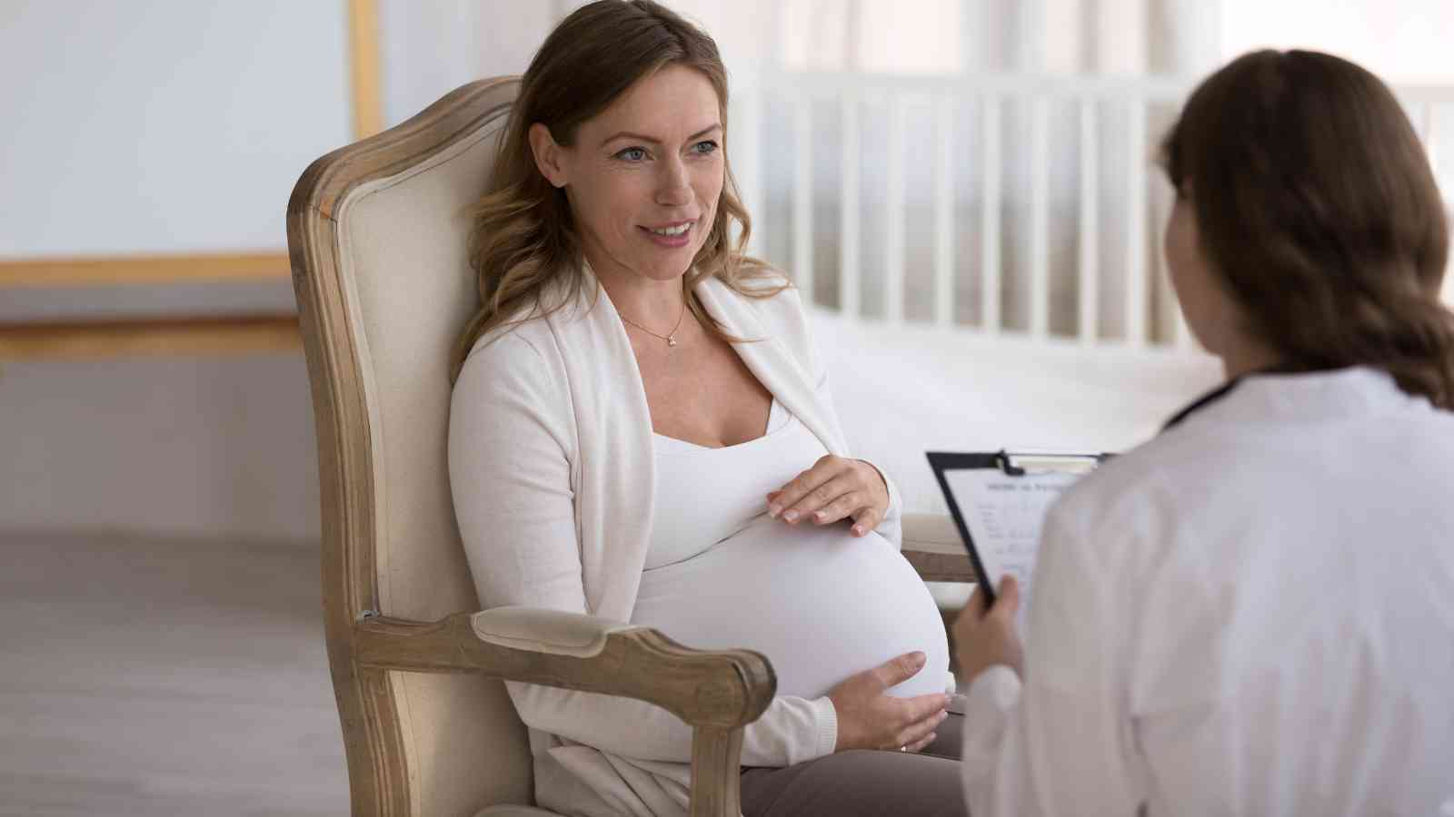 Pregnant woman sits in a chair, touching her belly, while talking to a health professional holding a clipboard.