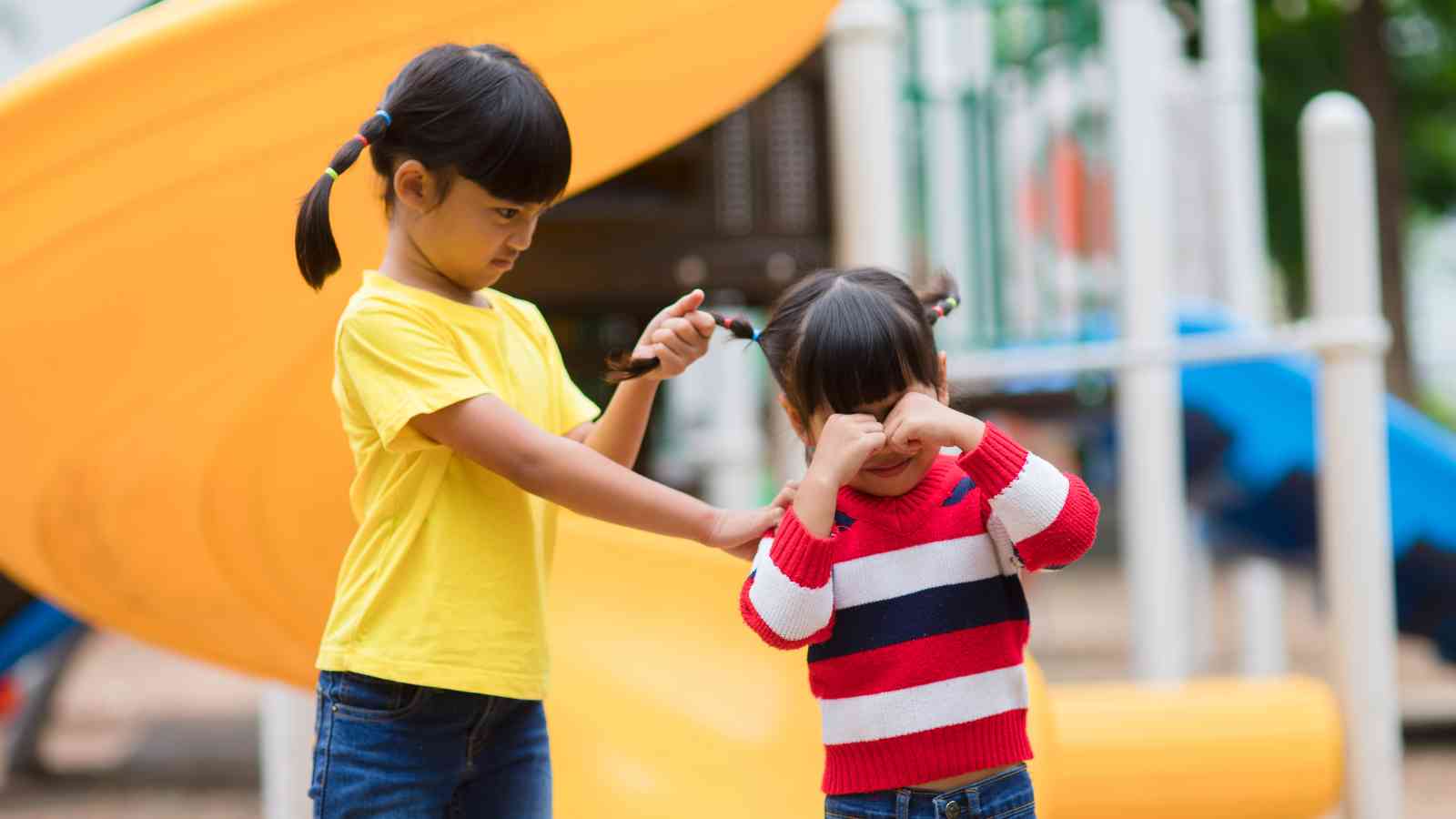 A child in a yellow shirt pulls another child's hair near a playground slide. The child in the striped shirt is crying.