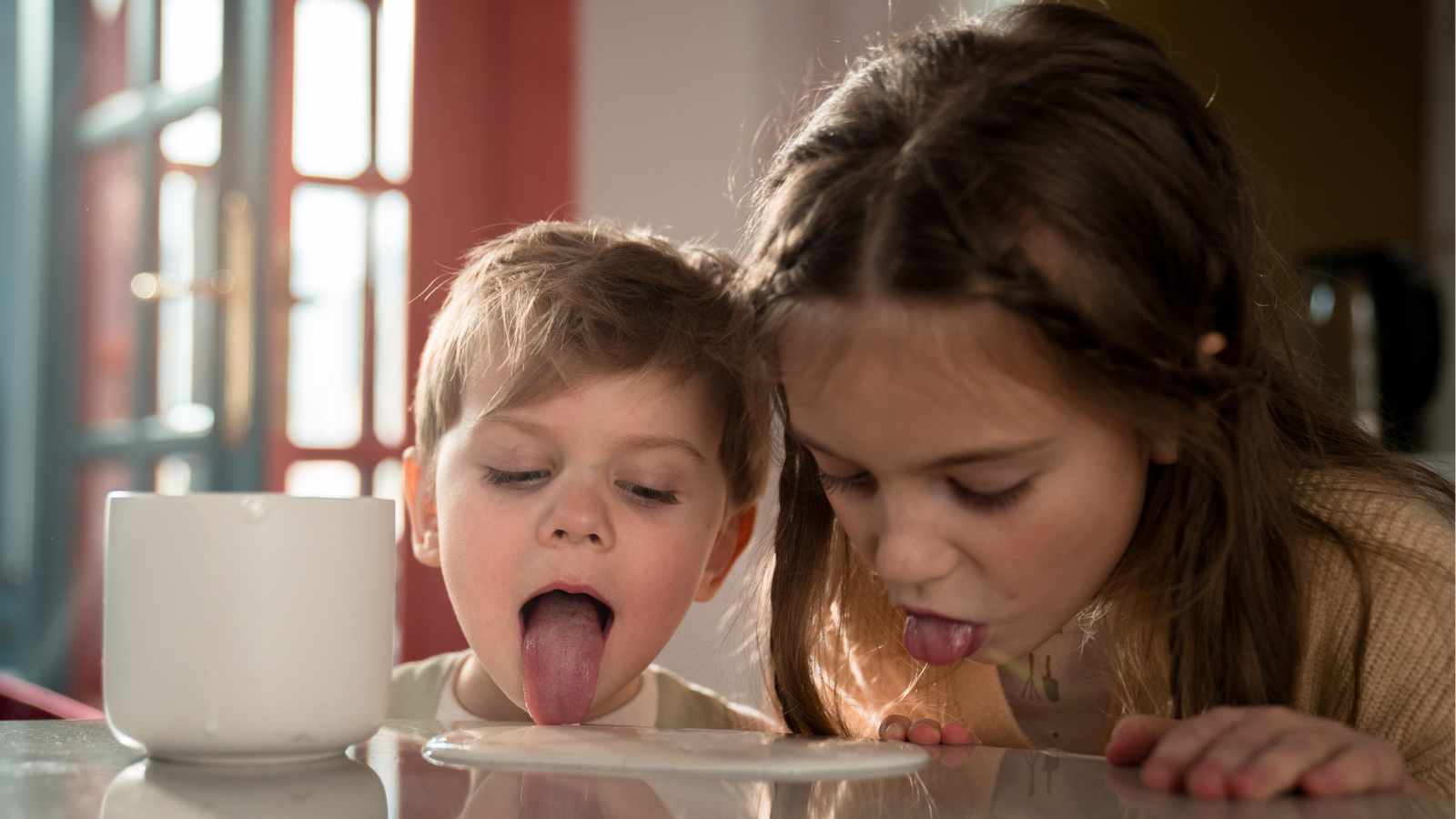 Two children with playful expressions lick milk spilled on a table next to a white mug.
