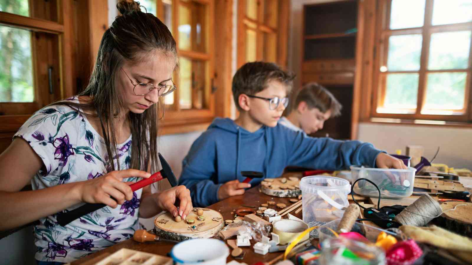 Three children focus on arts and crafts at a table filled with supplies in a well-lit room with wooden windows.