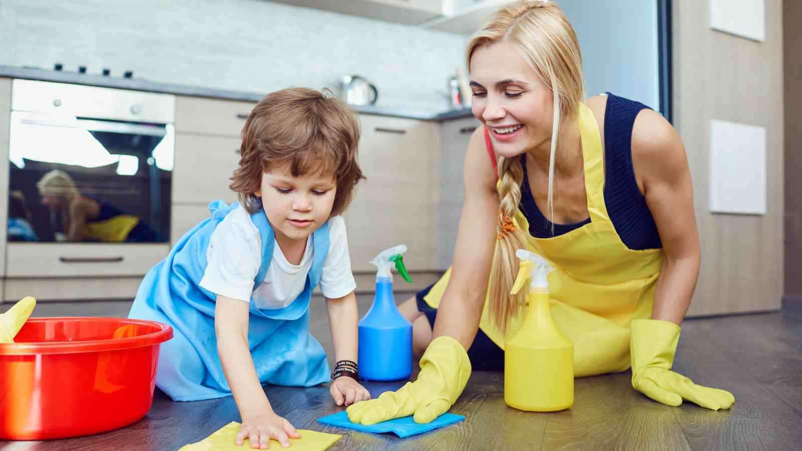 An adult and a child wearing aprons and gloves clean a kitchen floor with cleaning supplies and cloths on the ground.