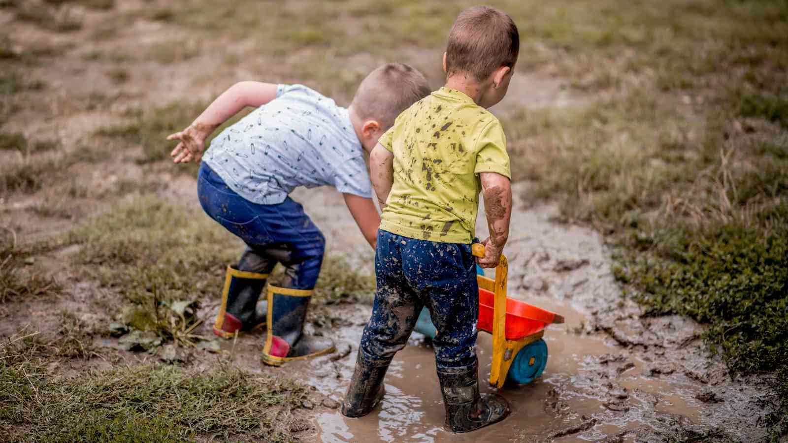 Two children in muddy clothes and boots play with a toy truck in a puddle on grassy ground.